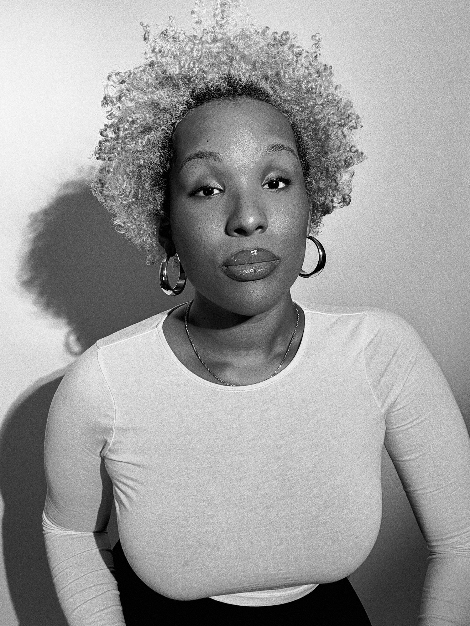 A woman with light curly hair wearing hoop earrings and a white long-sleeve top looks directly at the camera against a plain background. The photo is in black and white.